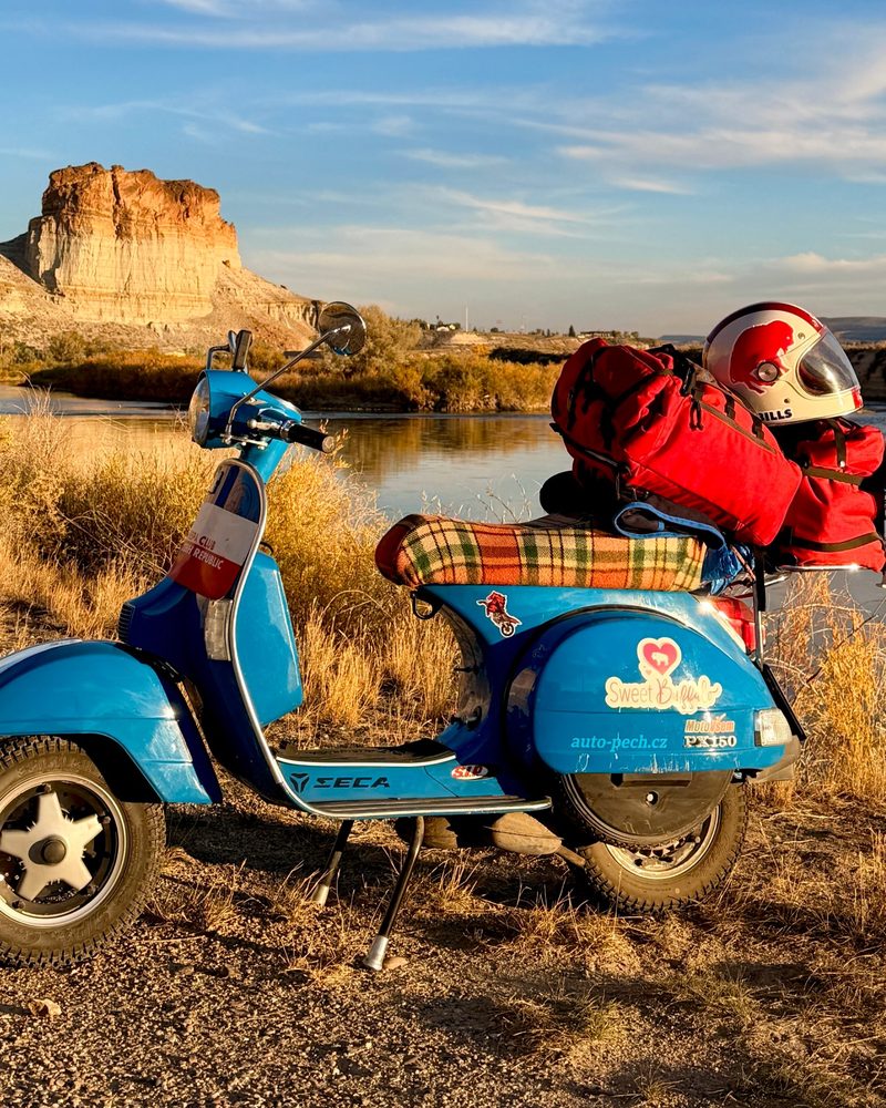 Adina, a blue 2011 Vespa loaded with red bags and a Bills helmet, parked by the Green River in Wyoming at sunset